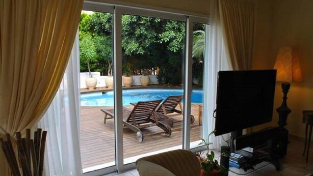 A room with a TV, a sofa, and a view of an outdoor swimming pool. - Paradise Villa Sounio (Saronikos)