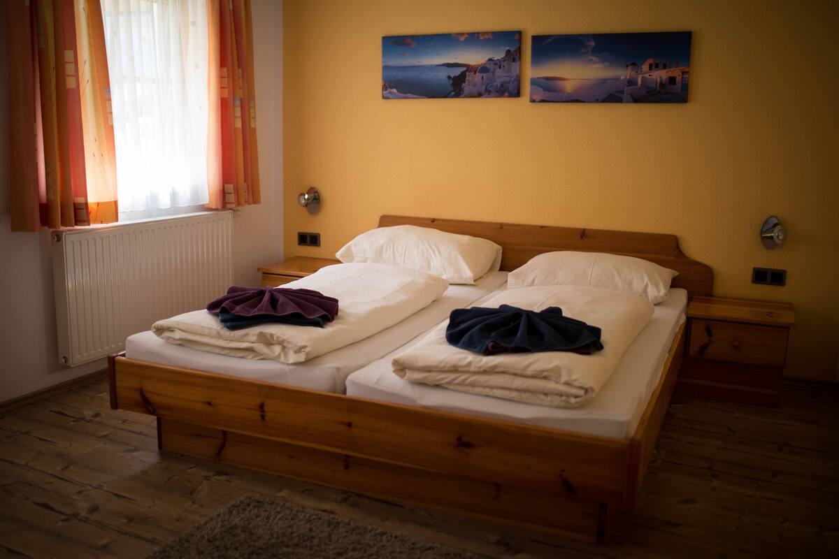 Uma cama de casal com duas almofadas e um cobertor, cabeceira de madeira, duas mesas de cabeceira, uma janela com cortinas e duas pinturas na parede.