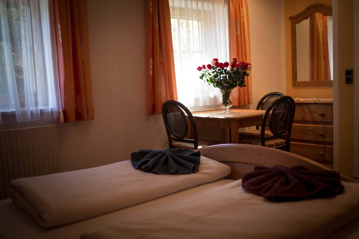 Quarto de hotel com cama, uma mesinha com um vaso de flores, duas cadeiras e um aparador.