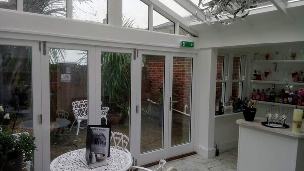A conservatory with a glass door leading to a patio area, white furniture, and a bar area with shelves of bottles. - Jireh House (Yarmouth)