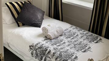 A single bed with a white and black floral bedspread, a white pillow, and a black pillow with a textured pattern. There is a window with curtains, a white door, and a small window with a view of the outside.