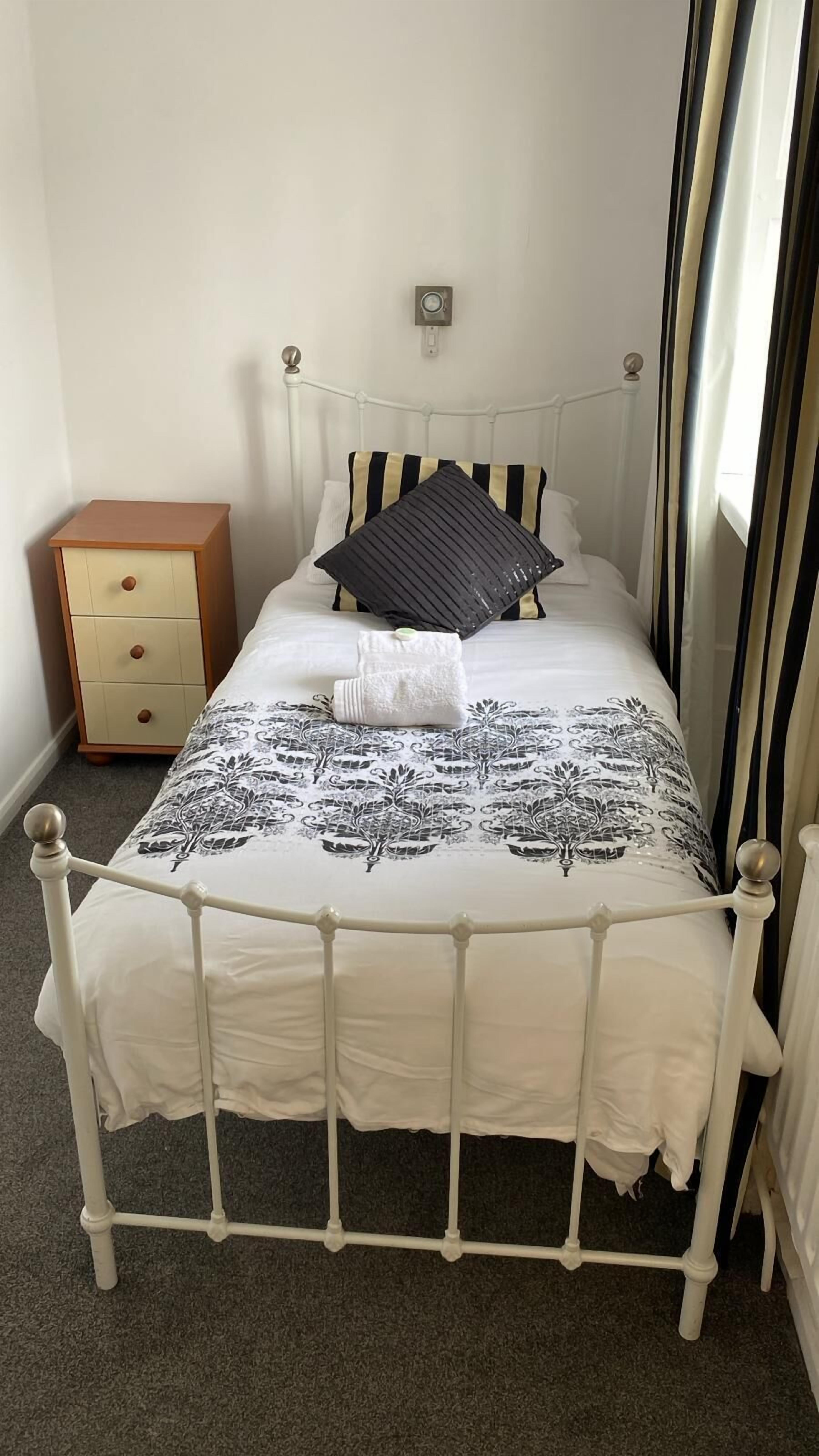 A single-bed room with a white metal frame, a patterned bedspread, two pillows, a bedside table, and a nightstand.