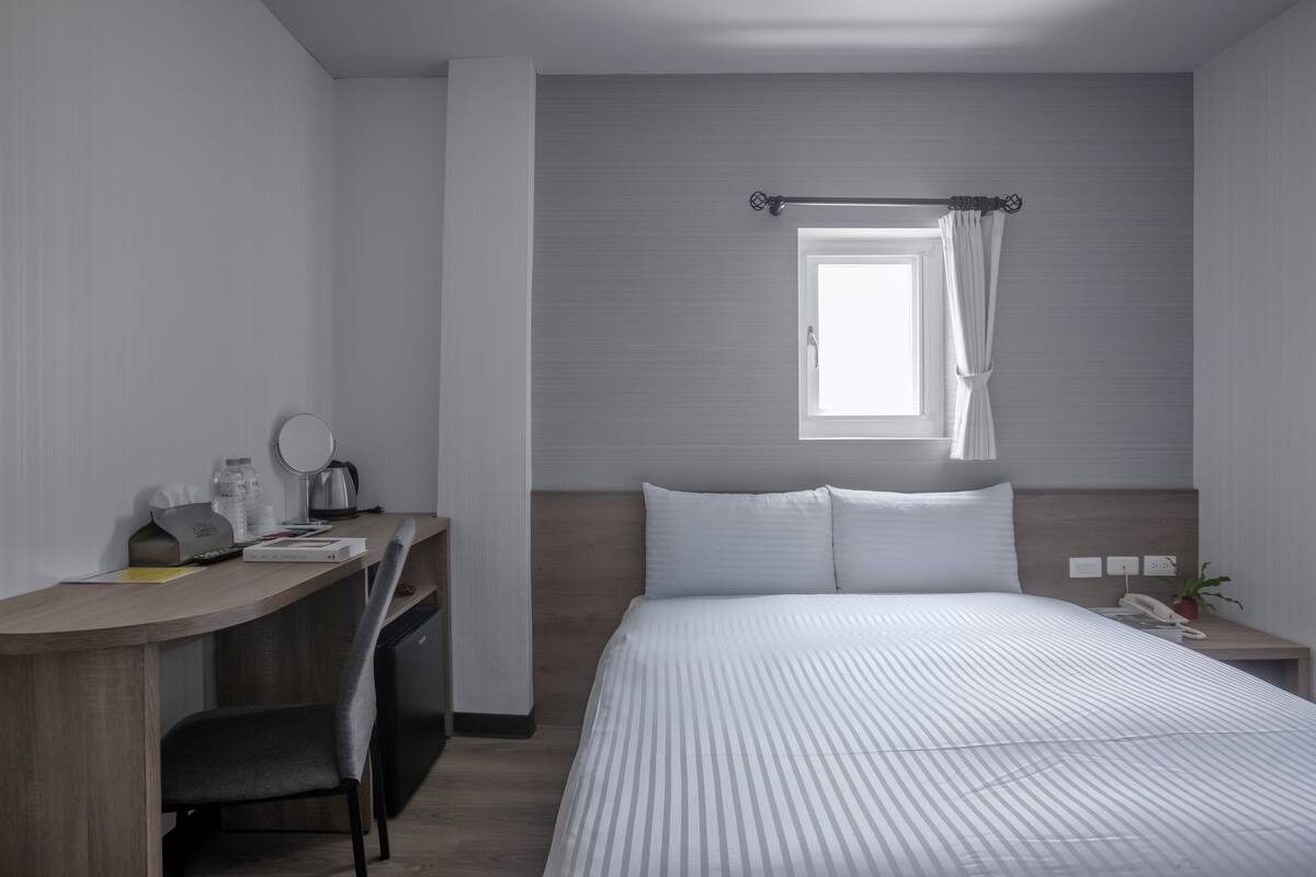Une chambre d’hôtel avec un lit, un bureau avec une chaise, une petite table avec un téléphone et une plante, et une fenêtre avec des rideaux.