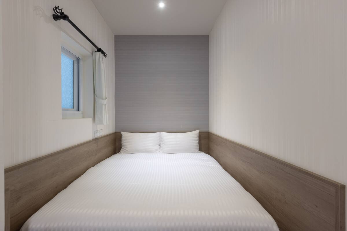 Une petite chambre avec un lit simple, une tête de lit en bois, une fenêtre avec un rideau et un mur de couleur claire.