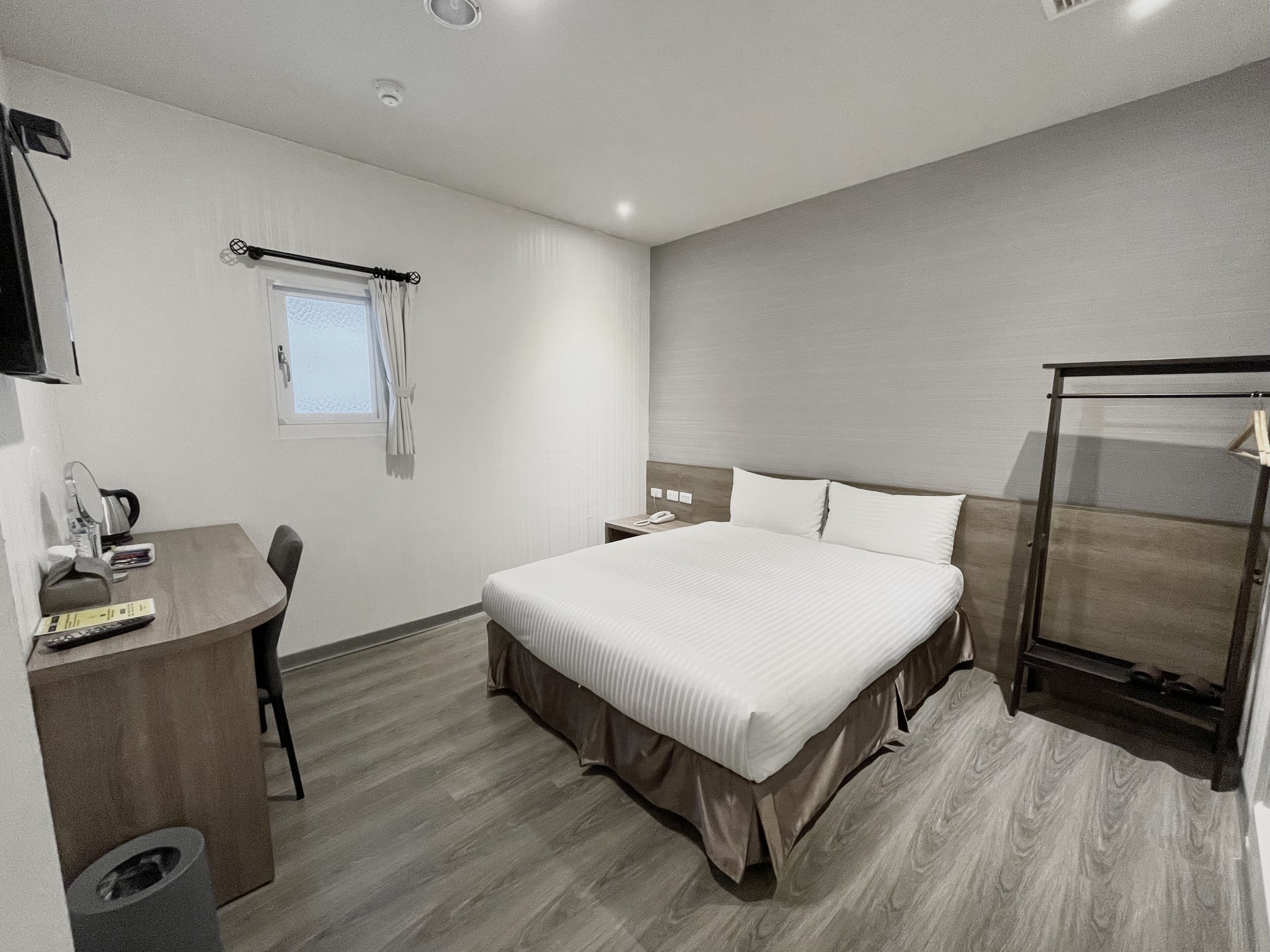 A modern hotel room with a large bed, a desk with a chair, a small window with curtains, and a bedside table with a lamp.