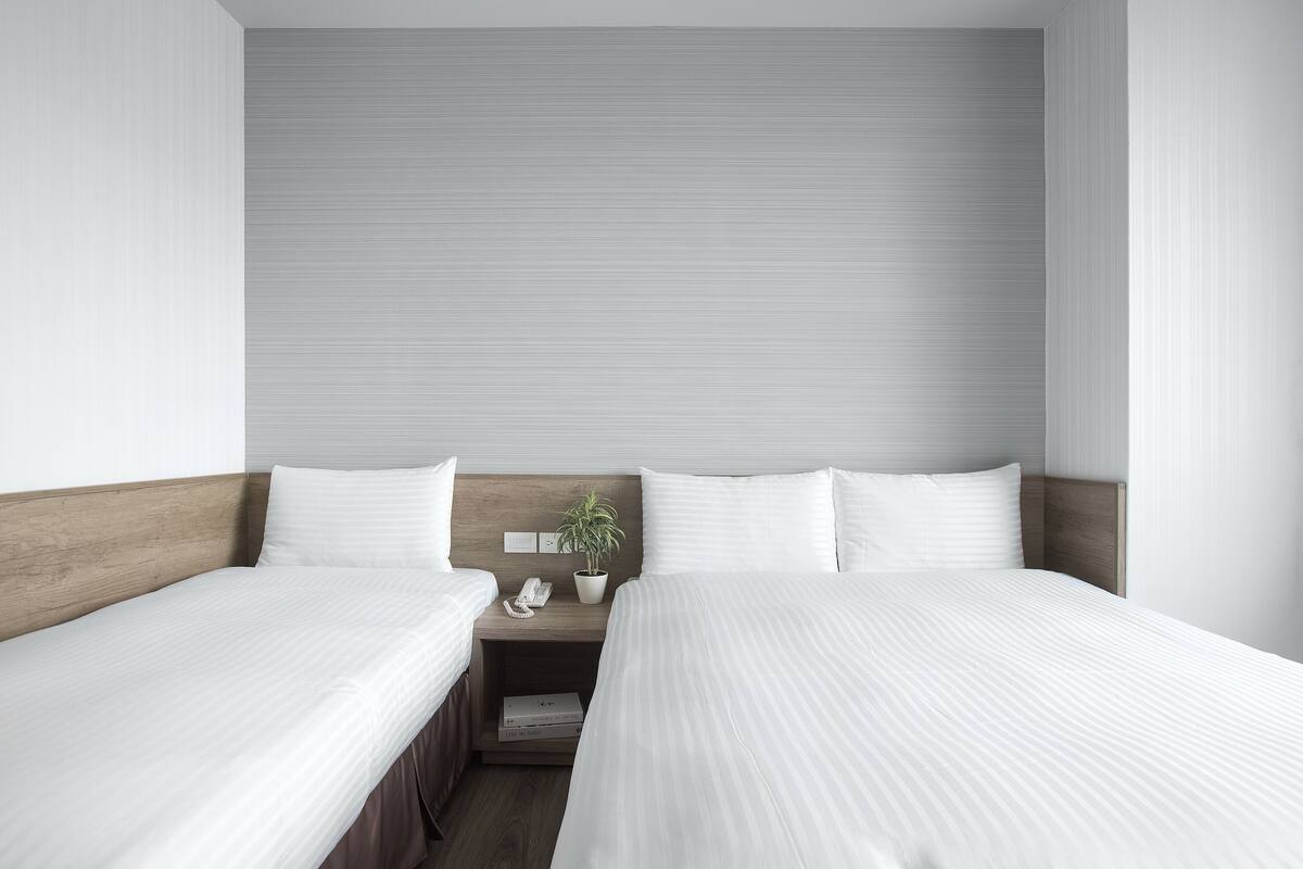 Une chambre d’hôtel avec deux lits simples, une tête de lit en bois, une table de chevet avec un téléphone et une plante, et un mur aux couleurs neutres.