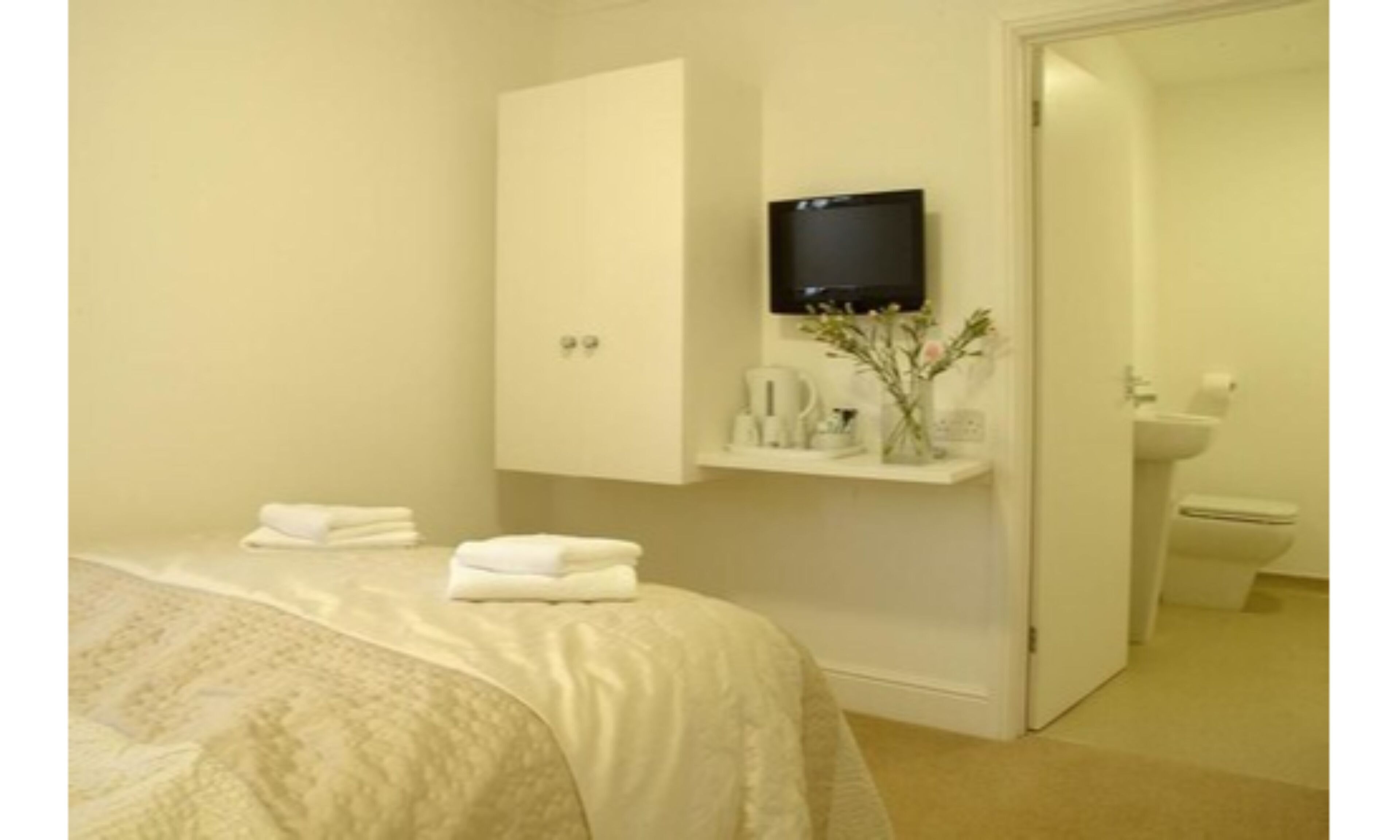 Habitación de hotel con cama, televisión montada en la pared, una pequeña estantería con artículos de aseo y un inodoro visible a través de una puerta abierta.