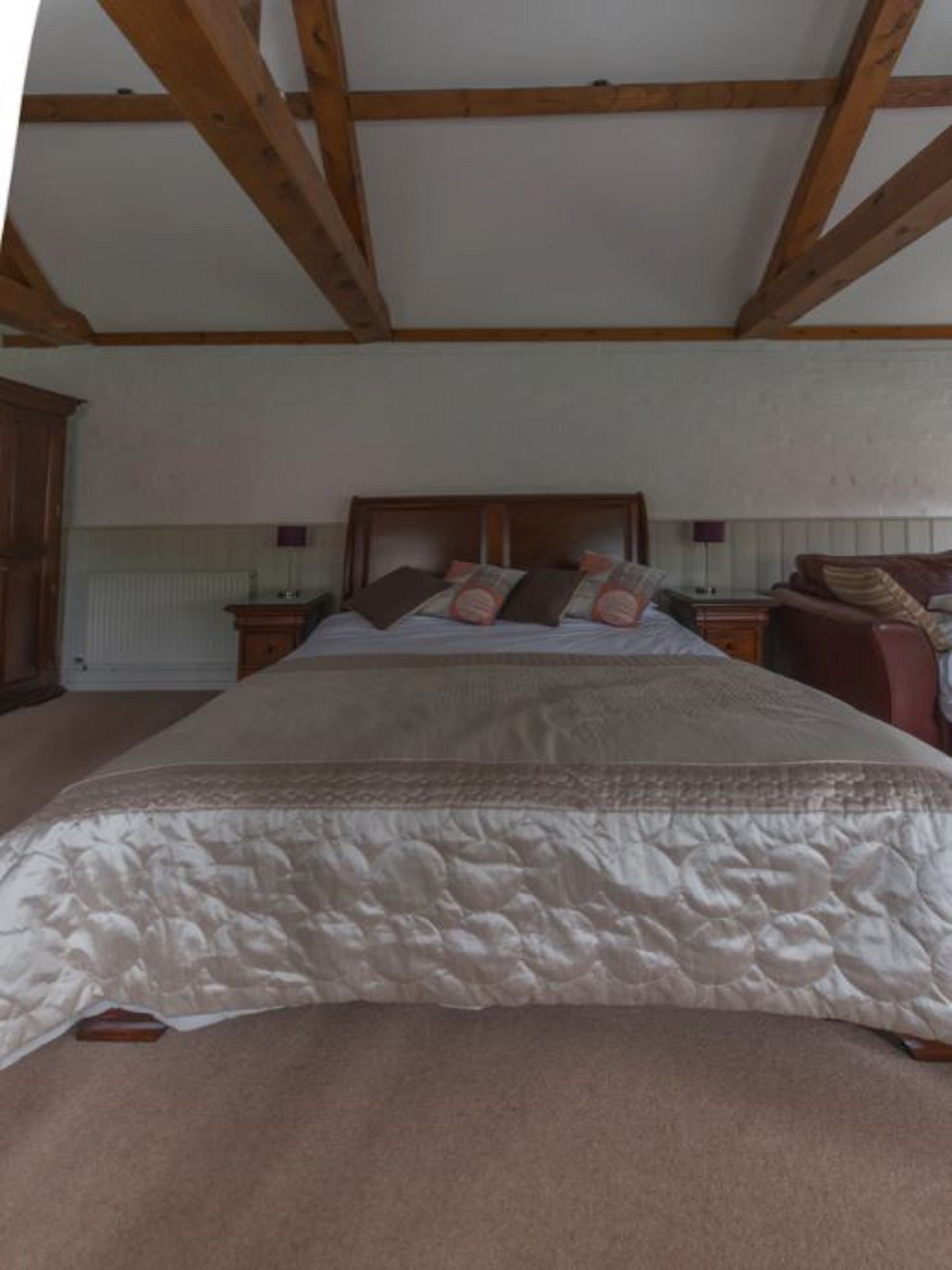 Ein Schlafzimmer mit Holzbalkendecke, einem Bett mit weißer Bettdecke und mehreren Kissen, einem Nachttisch mit Lampe und einem Holzschrank.