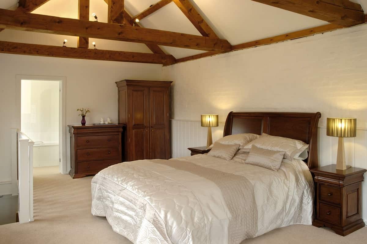 A bedroom with a large bed, wooden headboard, bedside tables with lamps, a wooden wardrobe, and a door leading to another room.