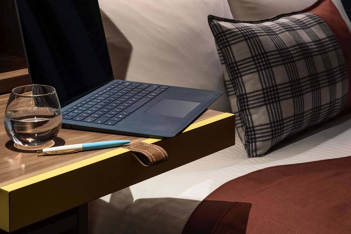 A modern hotel room with a laptop on a bedside table, a glass of water, and a pen.