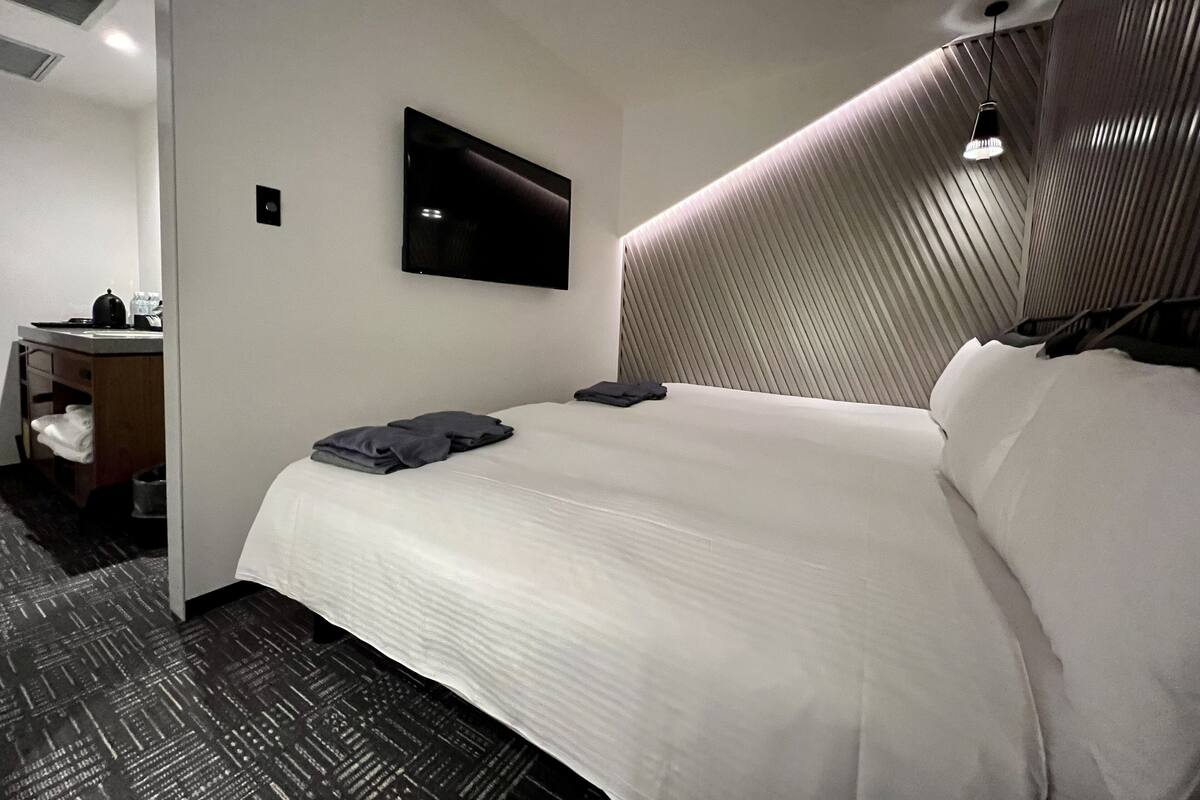 A hotel room with a neatly made bed, a flat-screen TV mounted on the wall, and a desk with a kettle and cups.