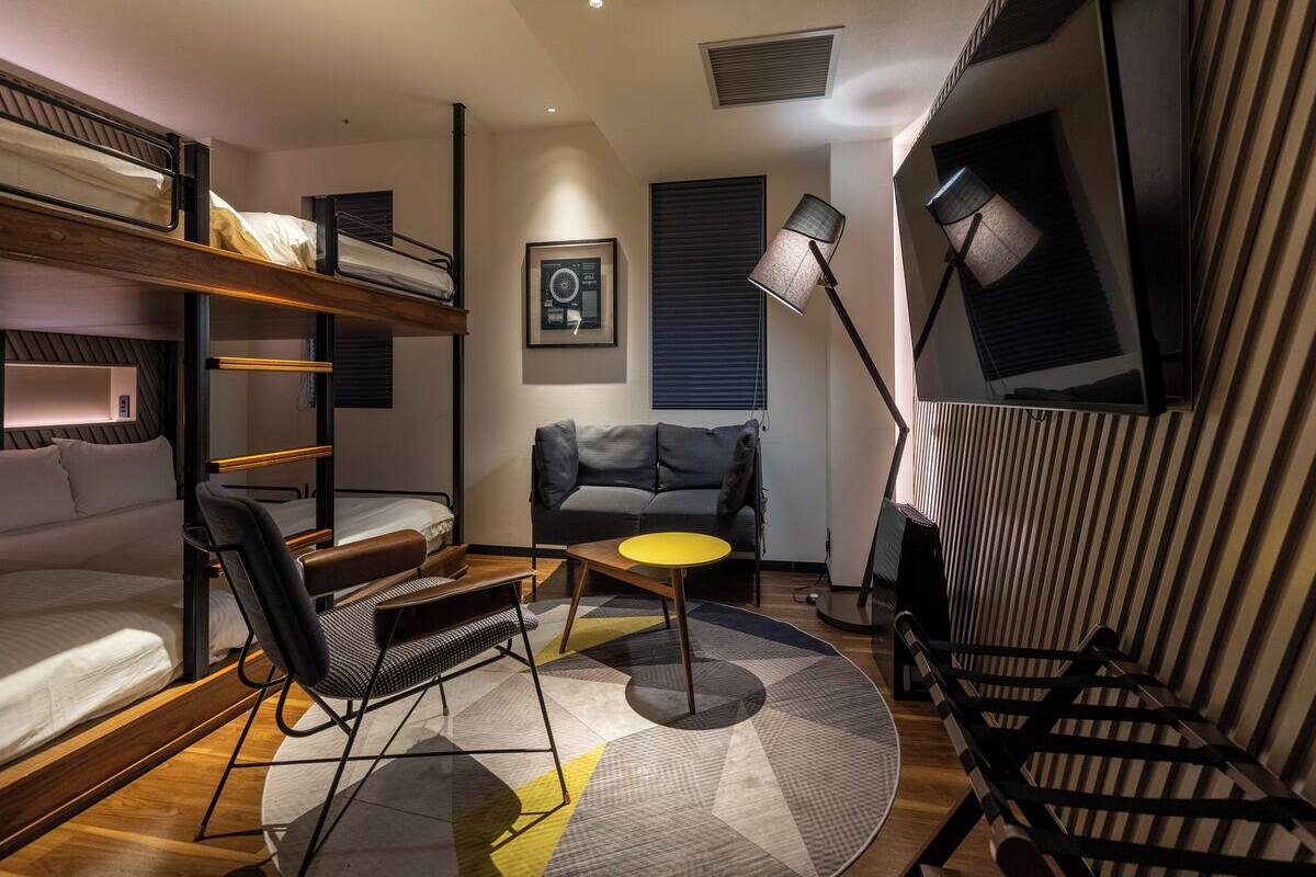 A compact hotel room with bunk beds, a sofa, a small table, and a floor lamp.
