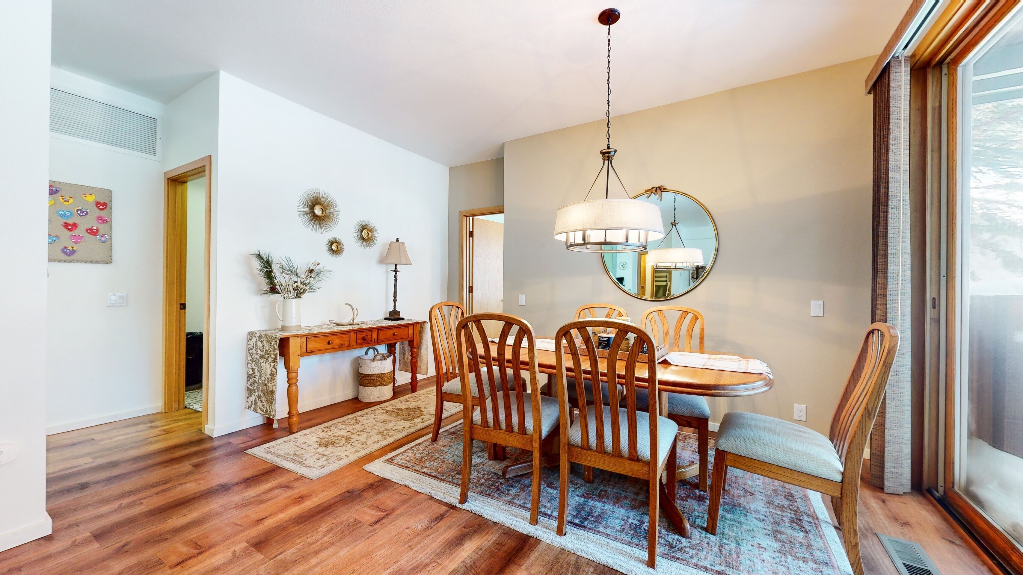 A dining area with a wooden table, four chairs, a chandelier, and a large mirror on the wall.