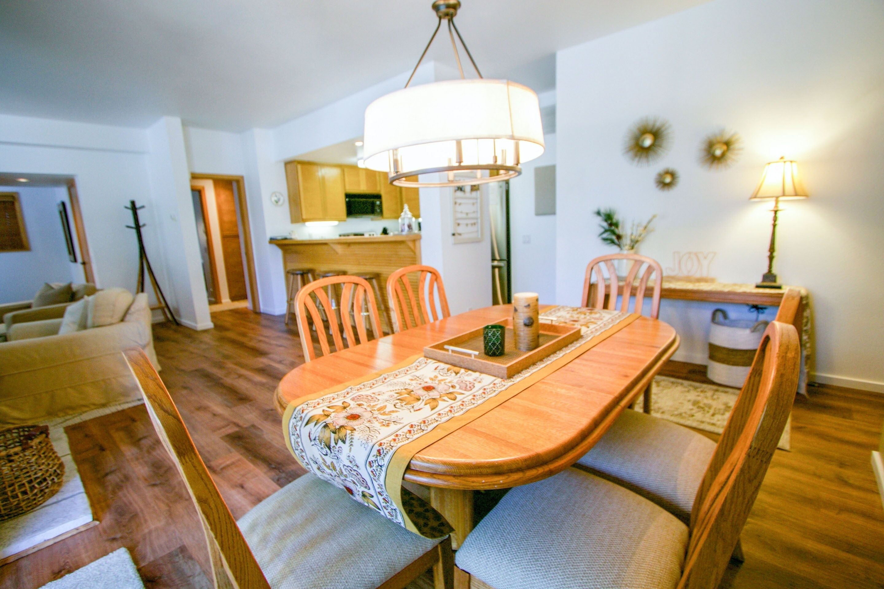 Un coin repas avec une table en bois, des chaises et un lustre. On aperçoit en arrière-plan une cuisine avec des meubles en bois et un réfrigérateur.