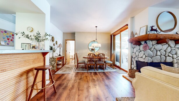 Un salón amplio con suelo de madera, una mesa de comedor con sillas, una zona de cocina con barra y una pared de piedra con chimenea.