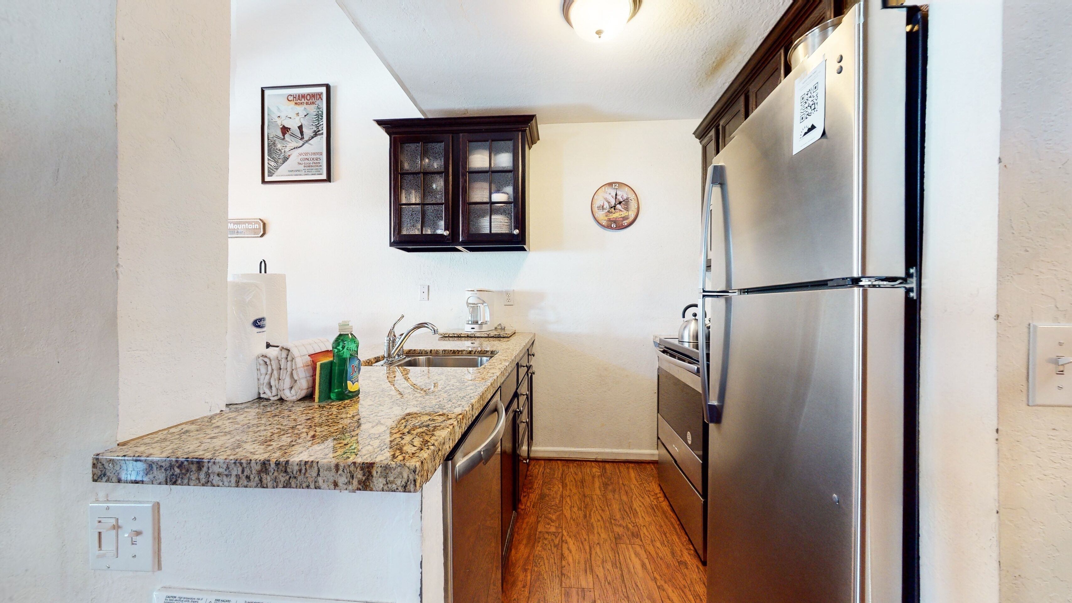 A compact kitchen with a stainless steel refrigerator, granite countertop, and a wooden floor.