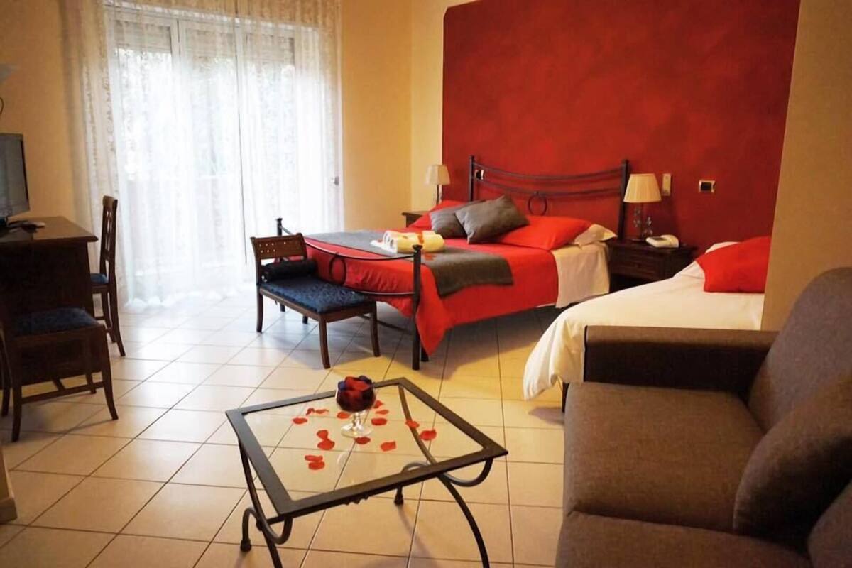 A hotel room with a bed, a desk, a chair, a glass-top table with a rose, and a red wall.