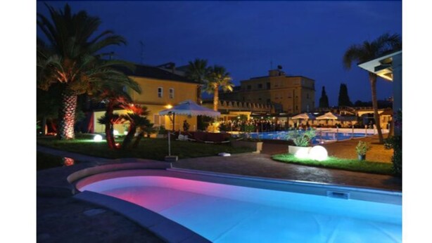 A swimming pool with a curved edge, surrounded by palm trees and buildings with warm lighting. - Park Hotel Paradiso (Piazza Armerina)