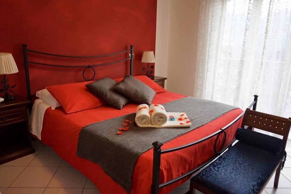 A bed with red bedding, a wooden headboard, a bench with a cushion, and two bedside lamps.