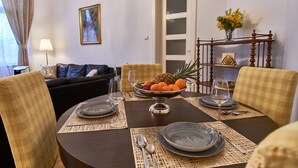 Comedor con una mesa redonda dispuesta para cuatro personas, mostrando un frutero, platos y cubiertos. Hay dos sillas con tapicería a cuadros, un sofá de color oscuro al fondo y un cuadro en la pared.