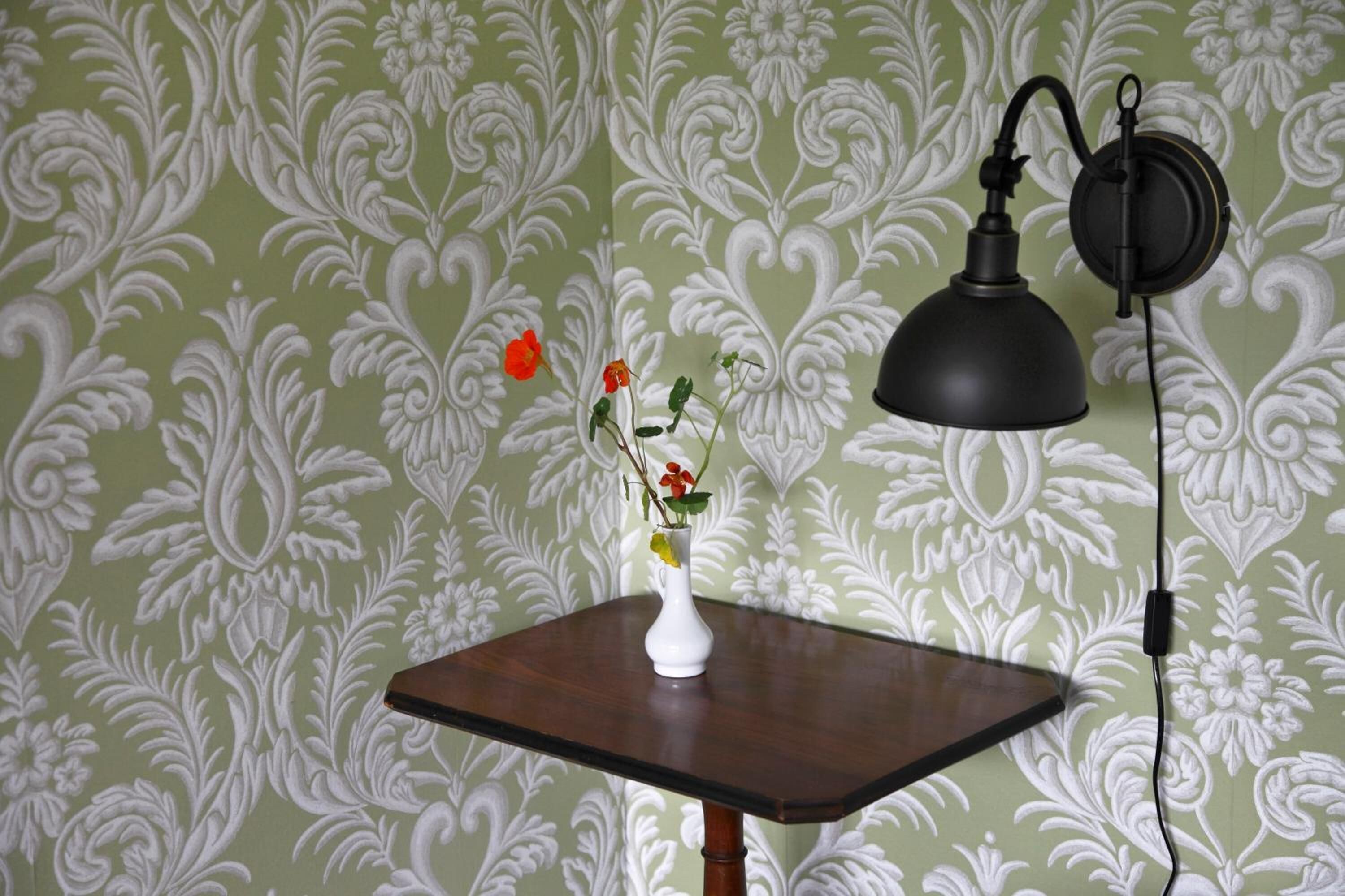 A table with a vase of flowers and a lamp against a patterned wallpaper background.