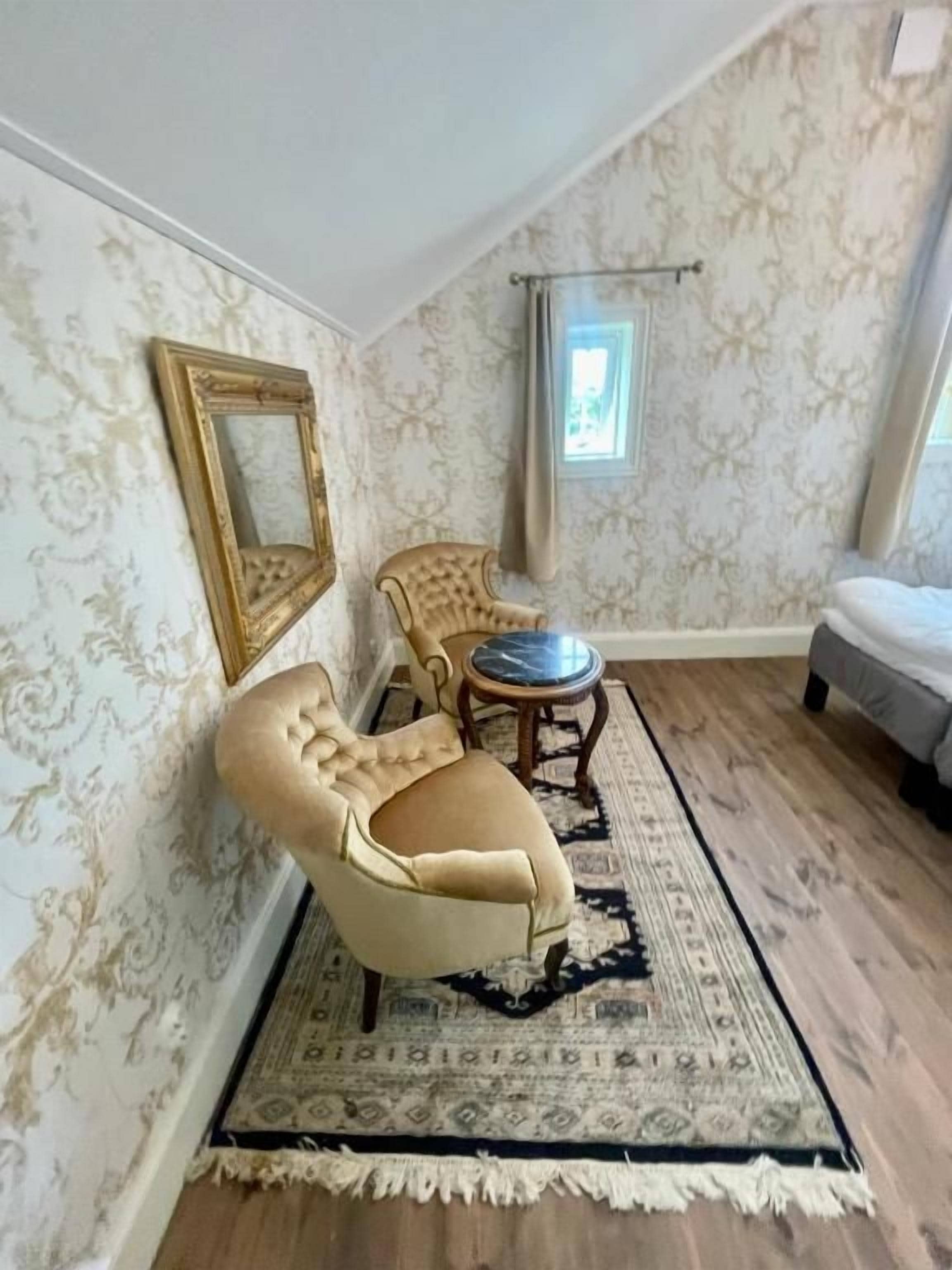 Una habitación con papel tapiz estampado, un espejo grande, un sillón capitoné, una mesa redonda y una cama.
