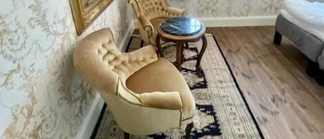 Una habitación con papel tapiz estampado, un espejo grande, un sillón capitoné, una mesa redonda y una cama.