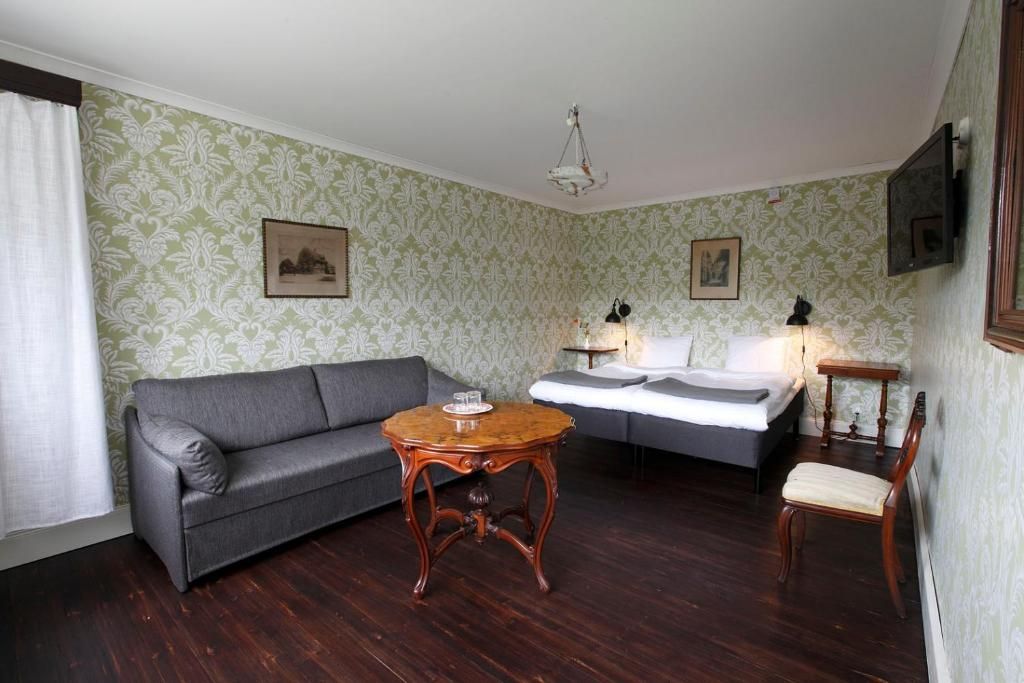 A room with a grey sofa, a small wooden table, a bed with white bedding, and a chair.