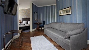 A room with a grey sofa, a small table, a TV mounted on the wall, a bed with a headboard, and a picture frame on the wall. - Ardrebo B&B (Ljugarn)