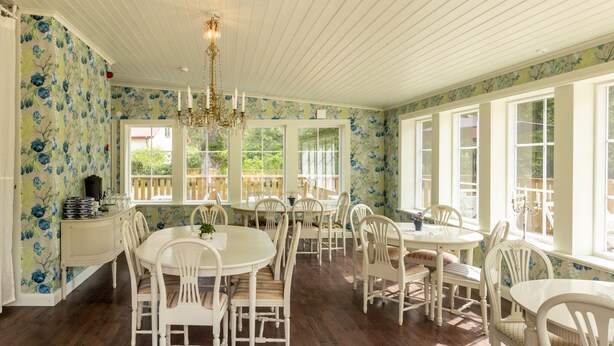 A dining area with white wooden tables and chairs, a chandelier, and large windows with a view of a deck. - Ardrebo B&B (Ljugarn)