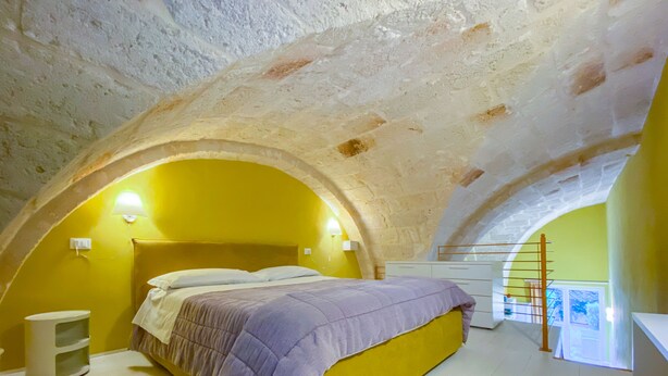 A bedroom with a high arched ceiling, a bed with a purple bedspread, a bedside table, and a nightstand. - Al Giardino Del Nespolo (Polignano a Mare)