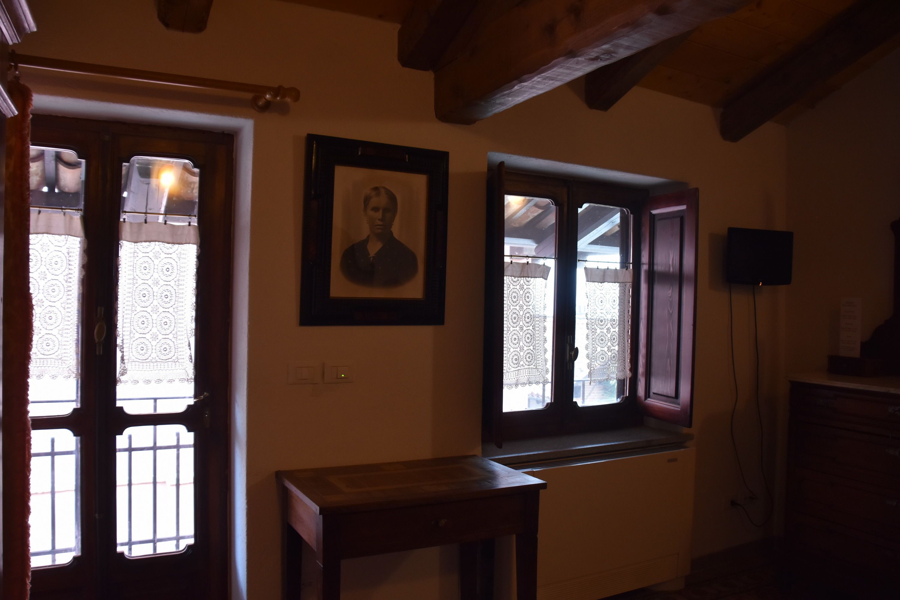 A room with a wooden ceiling, a framed portrait on the wall, and two windows with lace curtains.