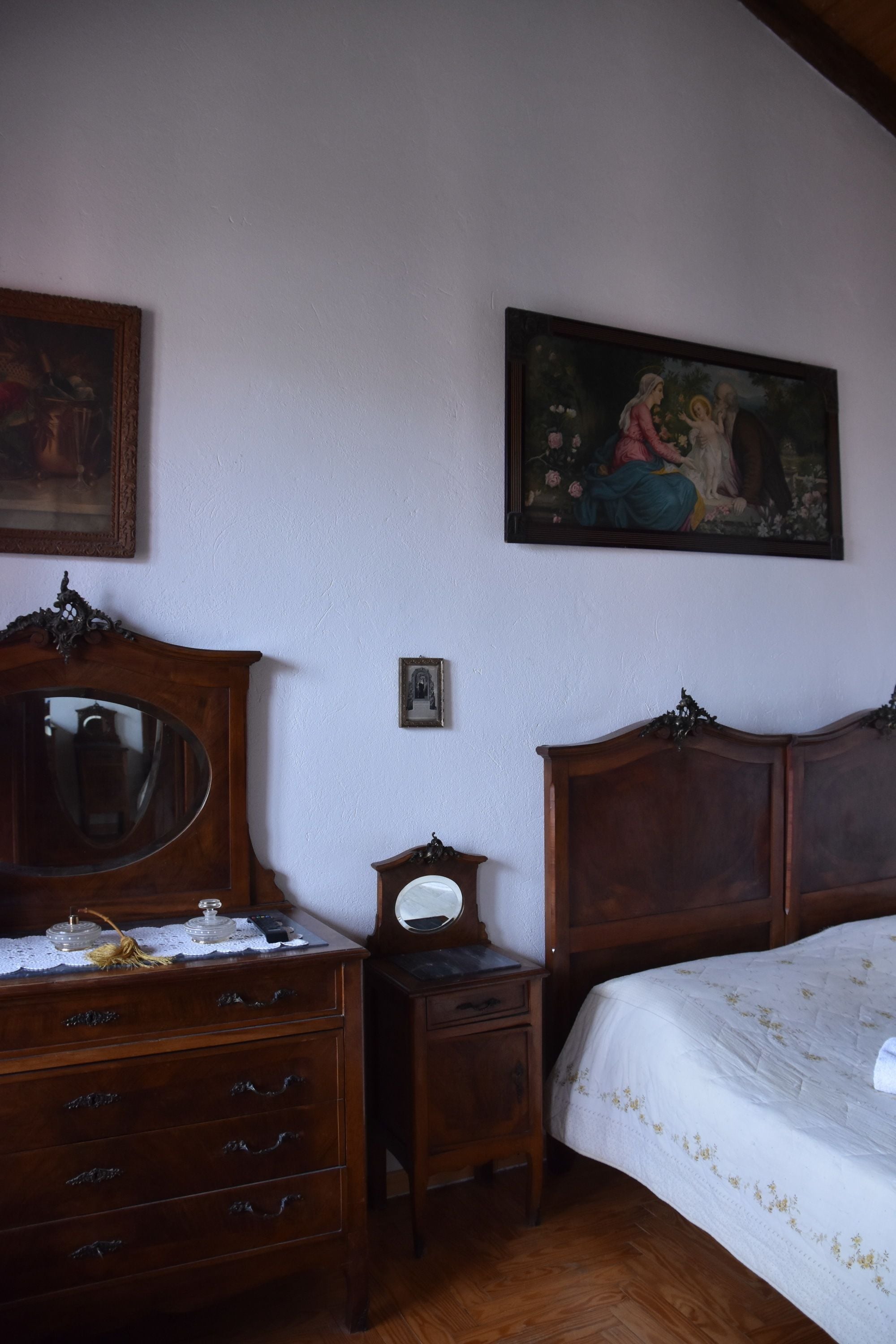 A bedroom with a wooden dresser, a bedside table, a bed with a floral bedspread, a mirror, and a painting on the wall.