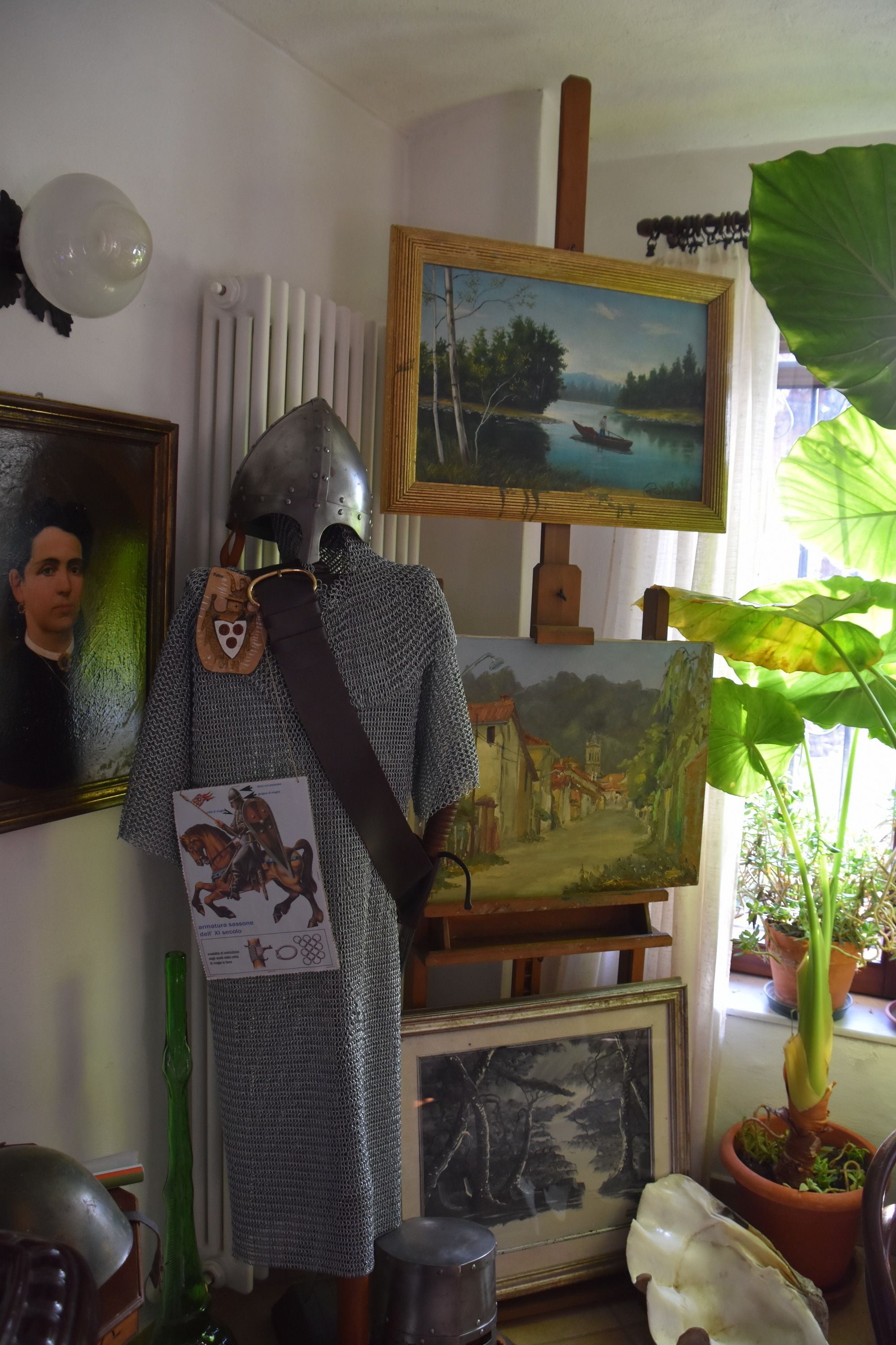Un maniquí vestido con armadura medieval, rodeado de varias pinturas y plantas en macetas.