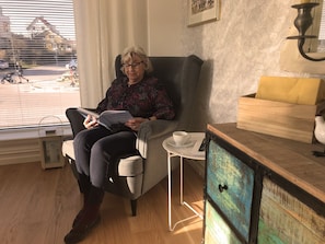 A person seated in a chair reading a book in a room with a window, a small table with a cup, and a wooden cabinet.