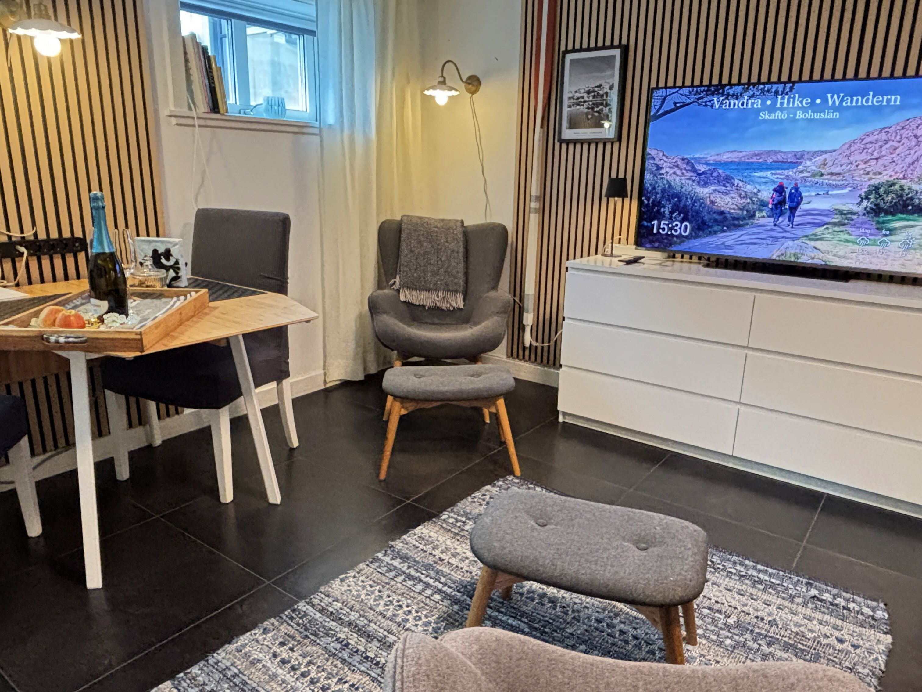 Ett modernt vardagsrum med ett matbord i trä, en grå fåtölj, en liten pall, en tv som visar ett vackert landskap och en mönstrad matta.