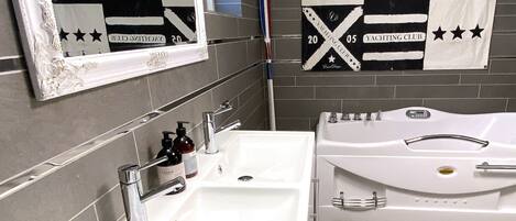 Une salle de bain avec un grand miroir, un lavabo et une baignoire. Des serviettes sont accrochées au mur, et une pancarte indique : « CLUB NAUTIQUE ».