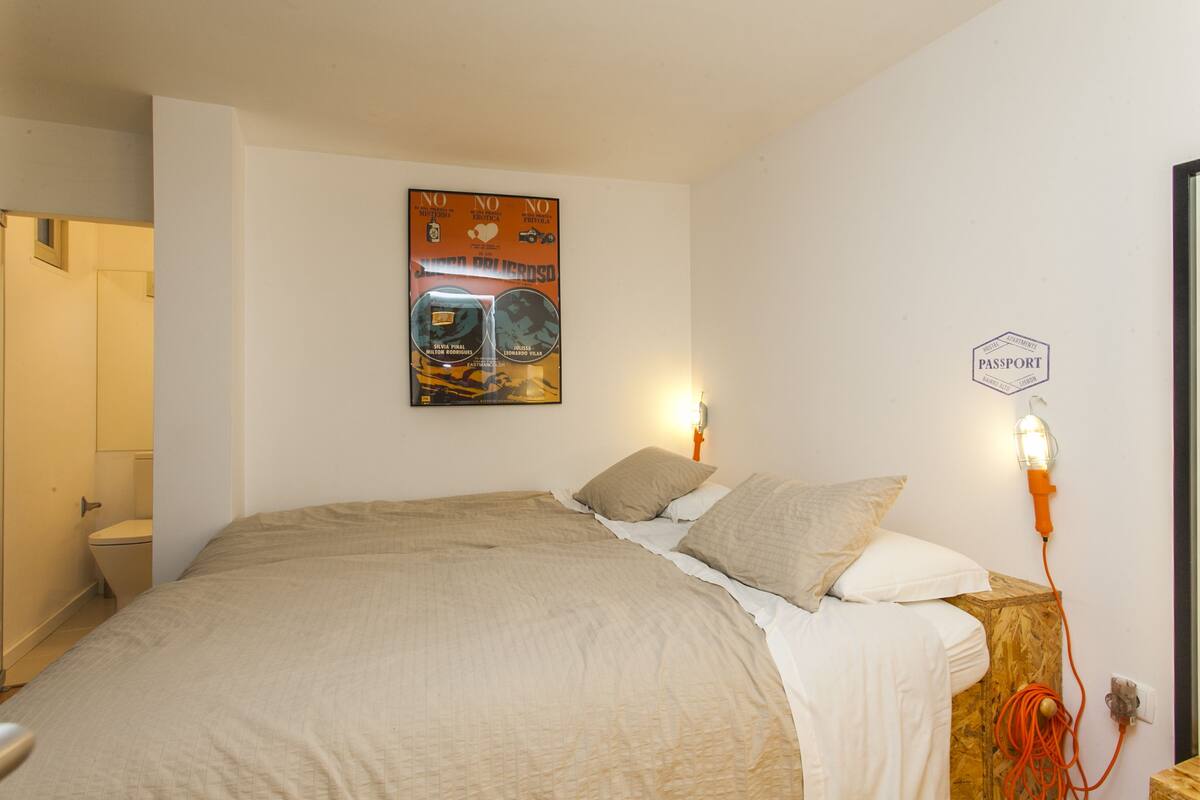Une chambre à coucher avec un lit, une affiche accrochée au mur, une table de chevet avec une lampe, et une porte sur le côté gauche.