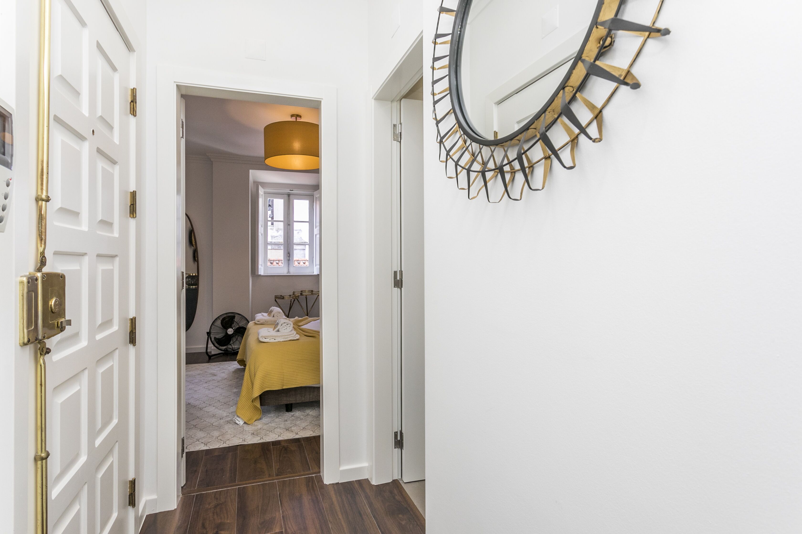 A hallway with a bed in the background, a round mirror on the wall, and a door with a brass handle.
