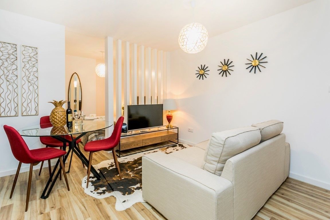 A modern living room with a glass dining table, red chairs, a beige sofa, a TV, and decorative wall art.