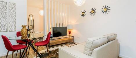 A modern living room with a glass dining table, red chairs, a beige sofa, a TV, and decorative wall art.