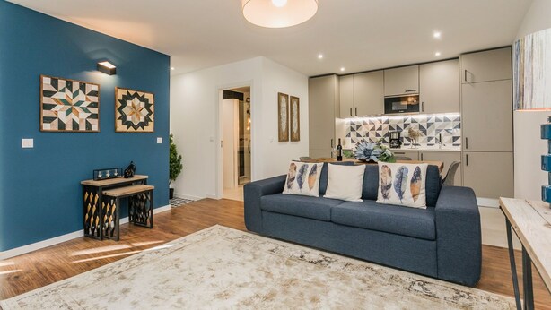 Een moderne woonkamer met een blauwe accentmuur, een grijze bank met decoratieve kussens, een houten salontafel en een groot vloerkleed.