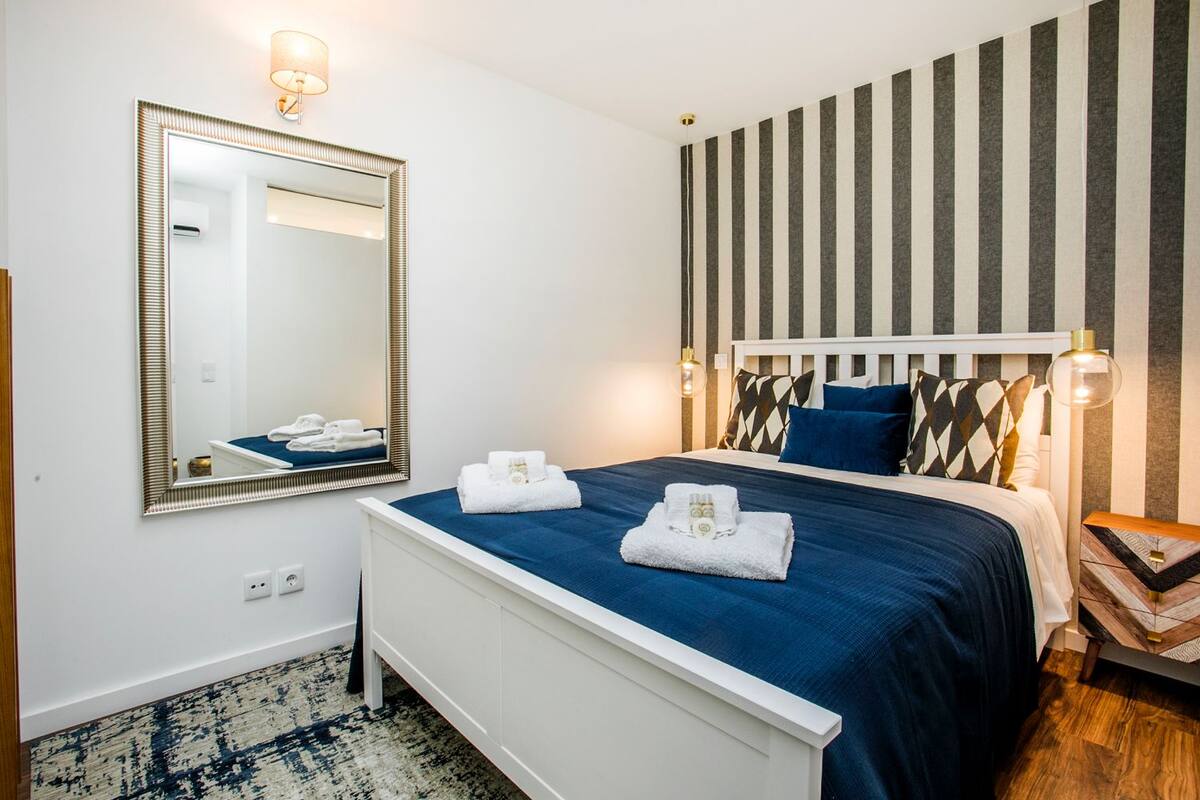 A hotel room with a large mirror, a bed with blue bedding, a striped wall, and a bedside table with a lamp.