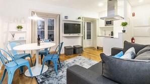 A modern living room with a grey sofa, a round table with blue chairs, a flat-screen TV, and a kitchen area with white cabinets and a microwave. - LxWay Apartments Galeria Alfama (Lisbon)