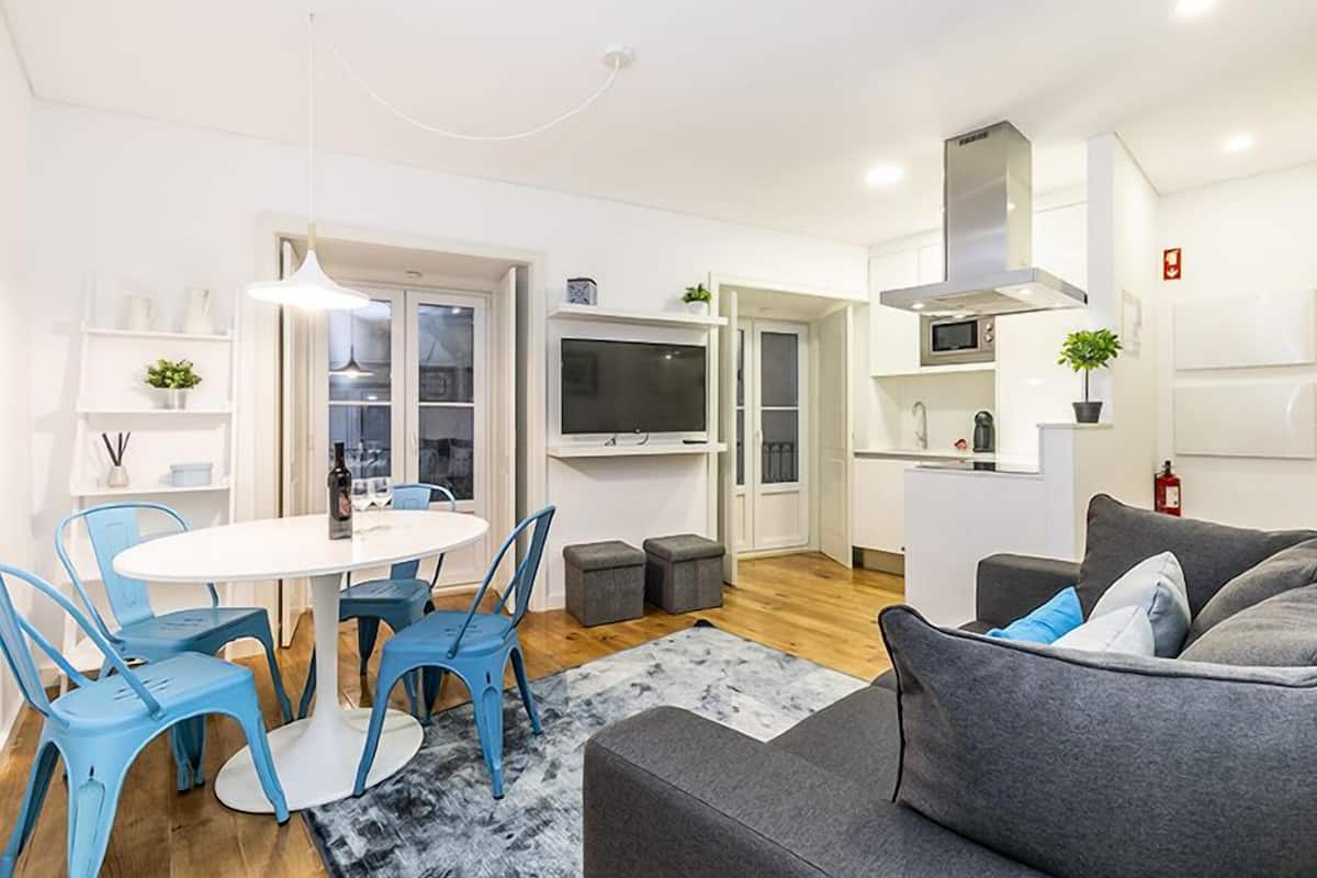 A modern living room with a grey sofa, a round table with blue chairs, a flat-screen TV, and a kitchen area with white cabinets and a microwave.