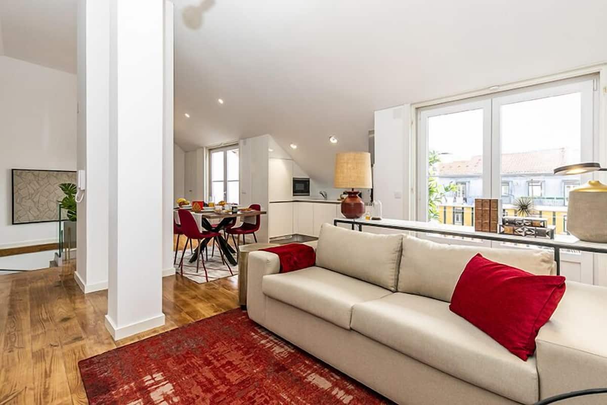 A modern living room with a sofa, red cushions, a dining area with chairs, and a kitchen in the background.