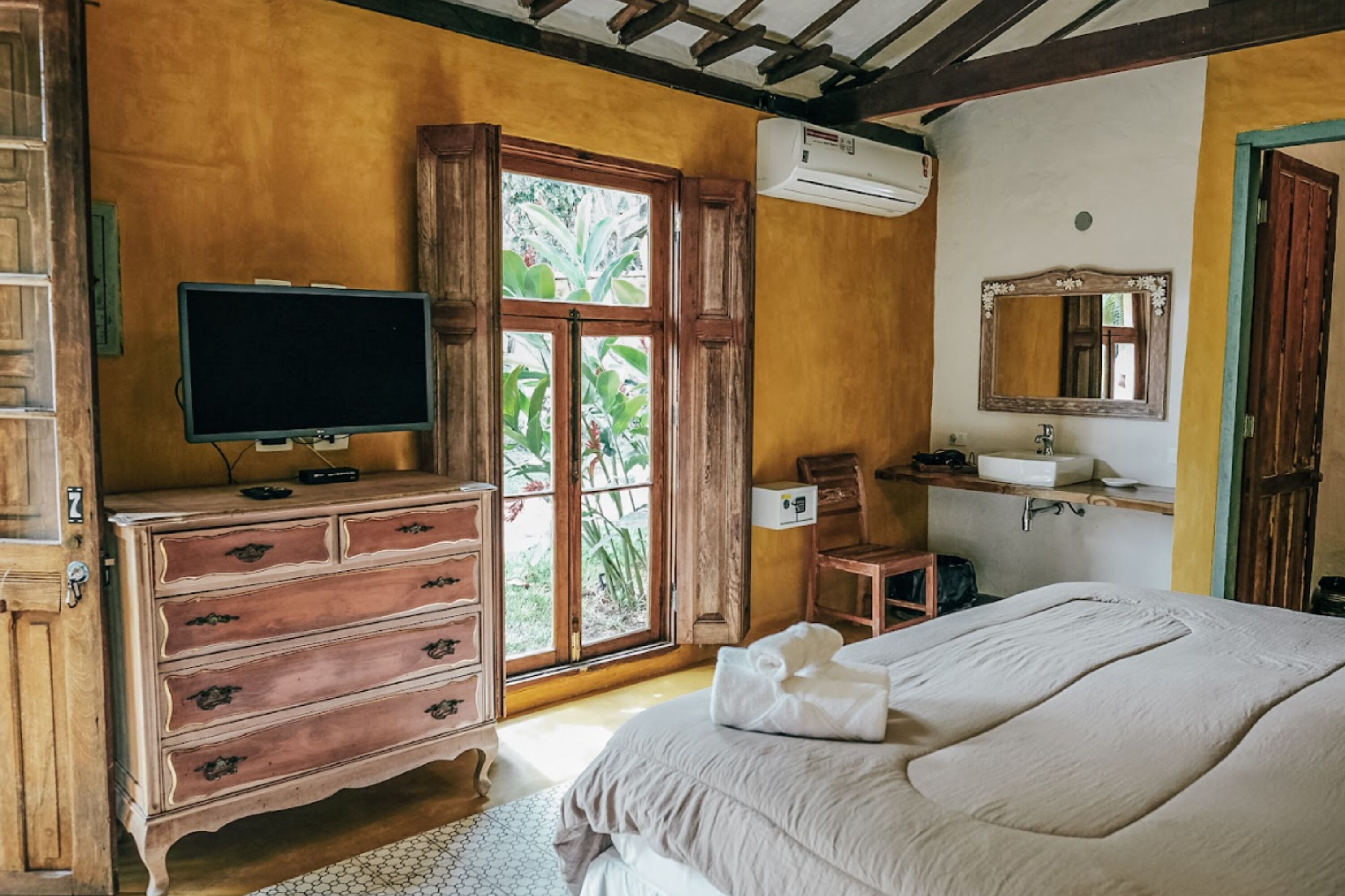 Ein Schlafzimmer mit Bett, Holzkommode, Fernseher, Fenster und Bad.