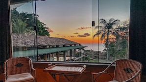 Un balcon avec des fauteuils en osier et une table, offrant une vue sur la côte, avec une construction à toit de chaume et des palmiers.