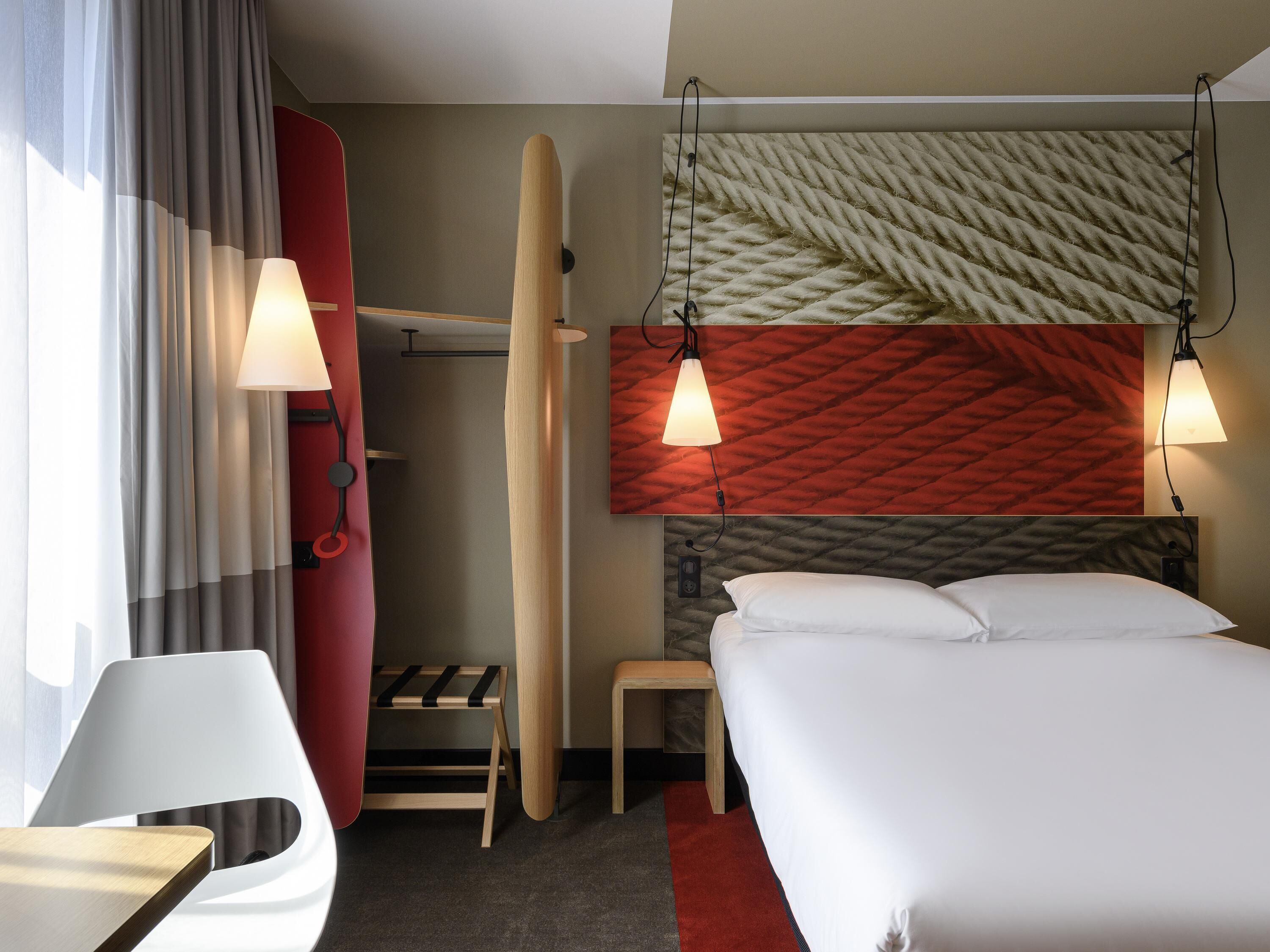 Habitación de hotel con cama, lámparas de noche, pared roja, cabecero de madera, una mesita, una silla y una ventana con cortinas.