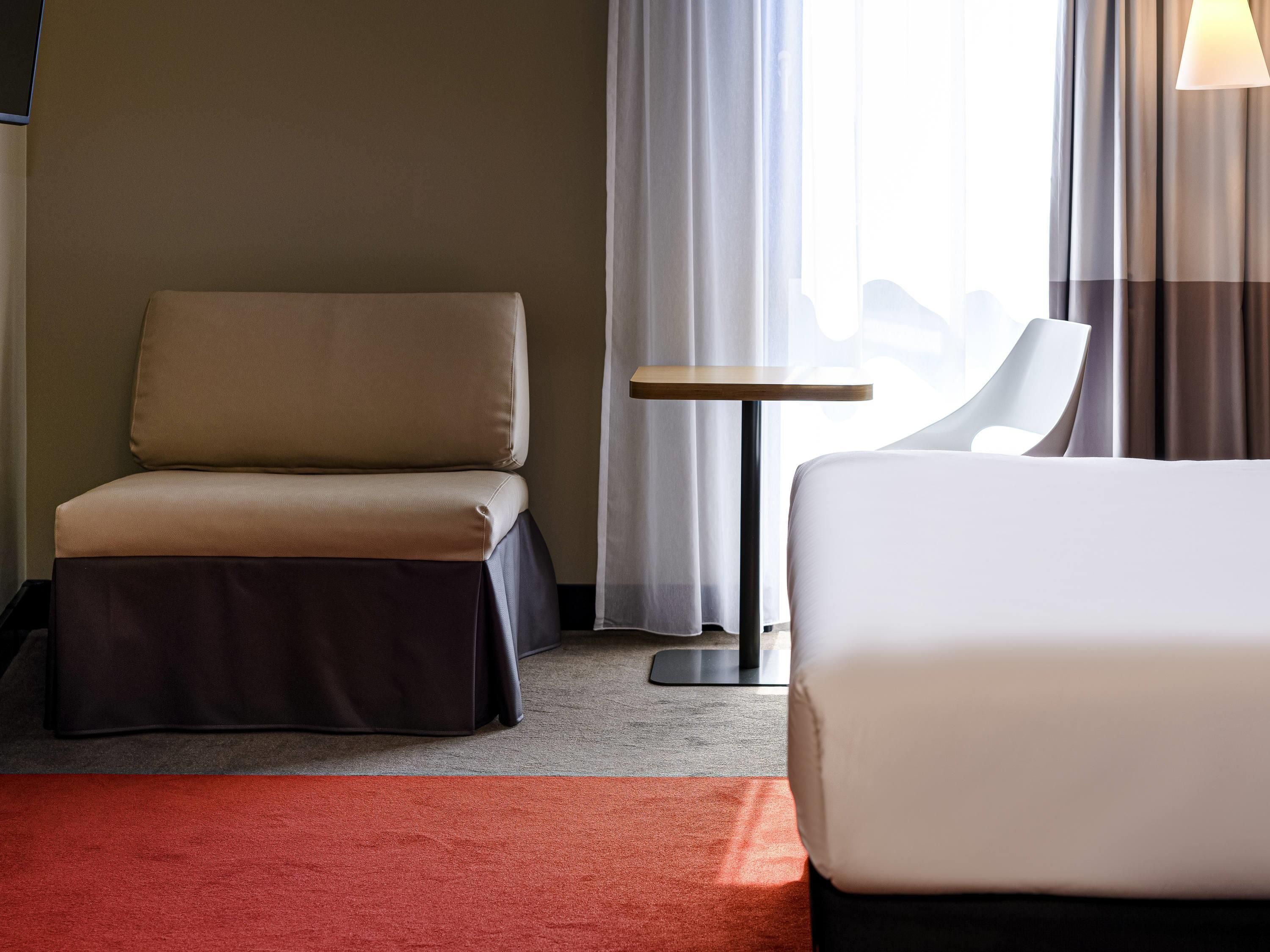 A hotel room with a bed, a sofa, a small table, and a view of the cityscape through the window.